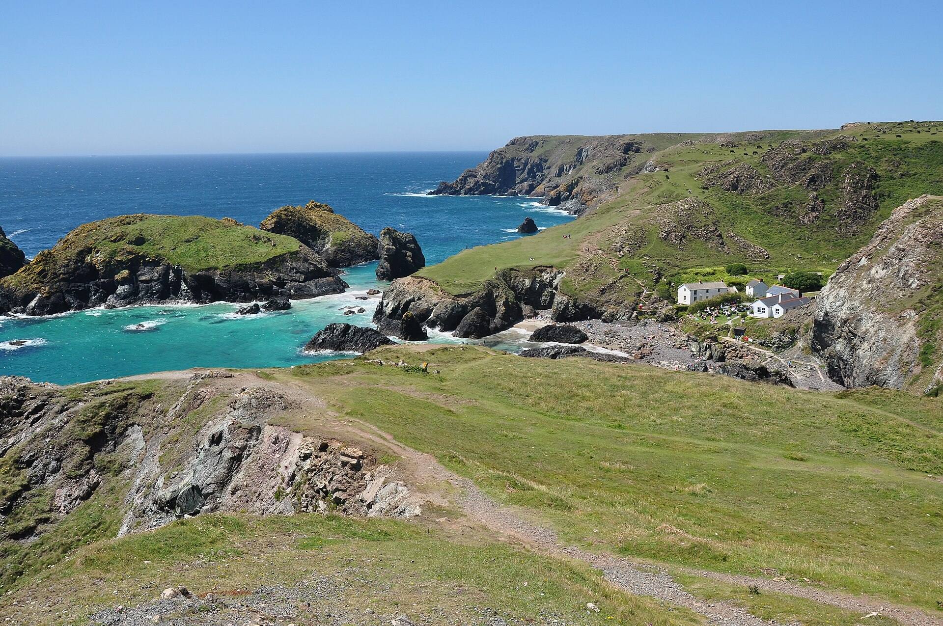 Kynance Cove from the east — turquoise water between serpentine rock stacks, green headlands, a white cottage tucked into the cliff.