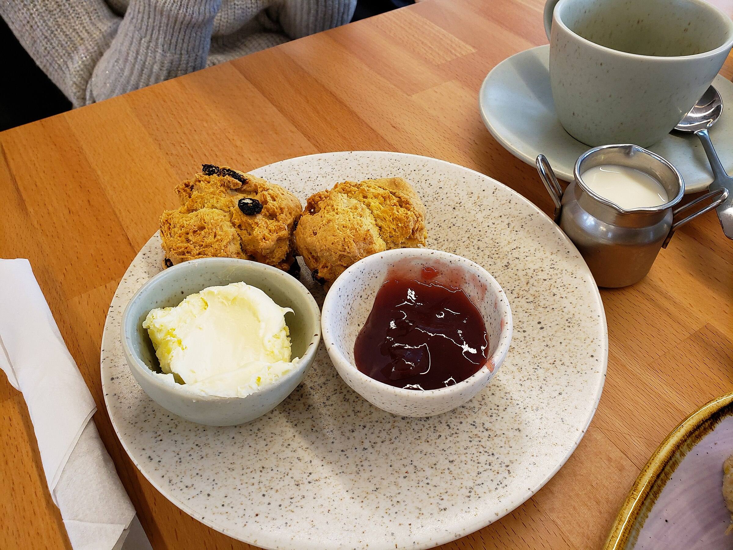 A Cornish cream tea — two scones with clotted cream and strawberry jam on a slate plate with a pot of tea behind.