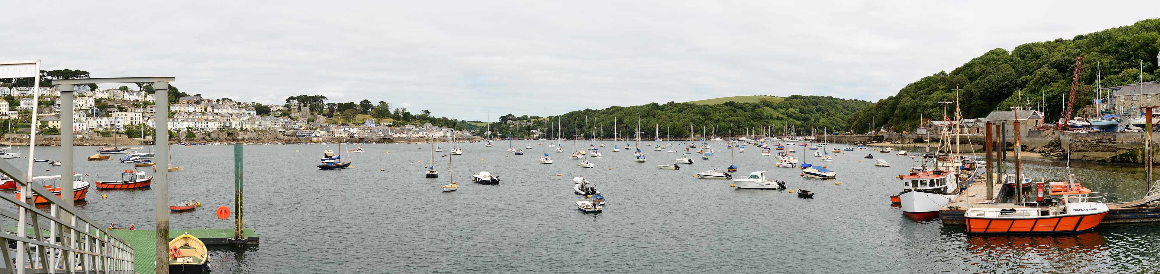 The Fowey estuary in soft afternoon light — terraced houses climbing the hillside, boats moored on the river, woods on the opposite bank.
