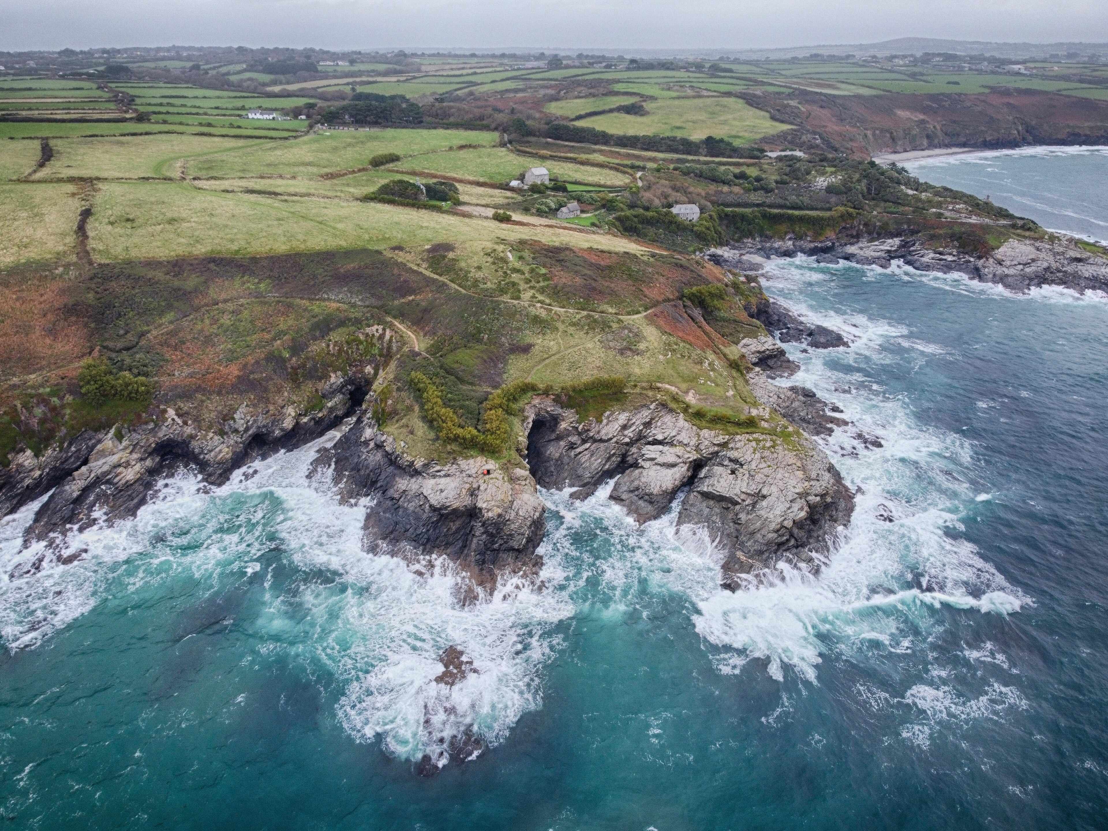Aerial view of Prussia Cove — waves breaking against rugged granite cliffs where green Cornish fields meet turquoise sea.