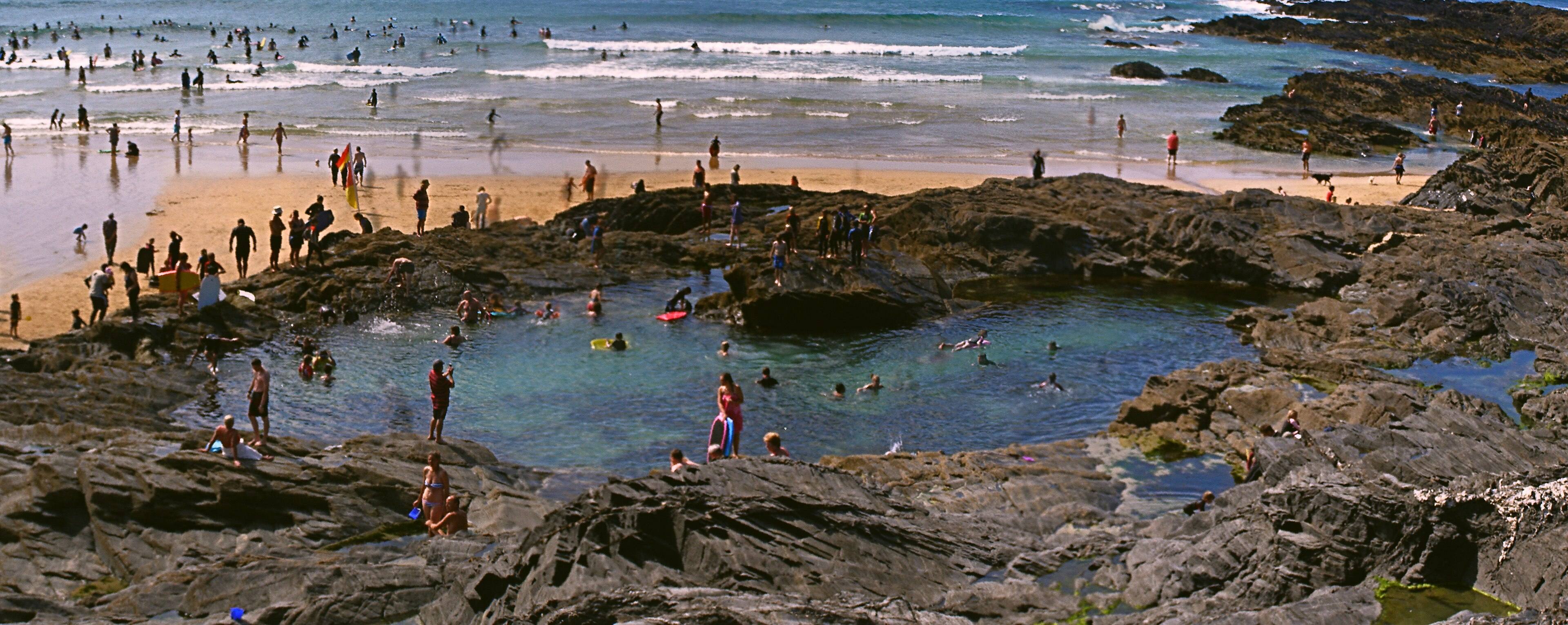 A child crouched at a Cornish rock pool with a small net — granite boulders, brown seaweed and a shallow pool of clear water teeming with shrimp and tiny crabs.